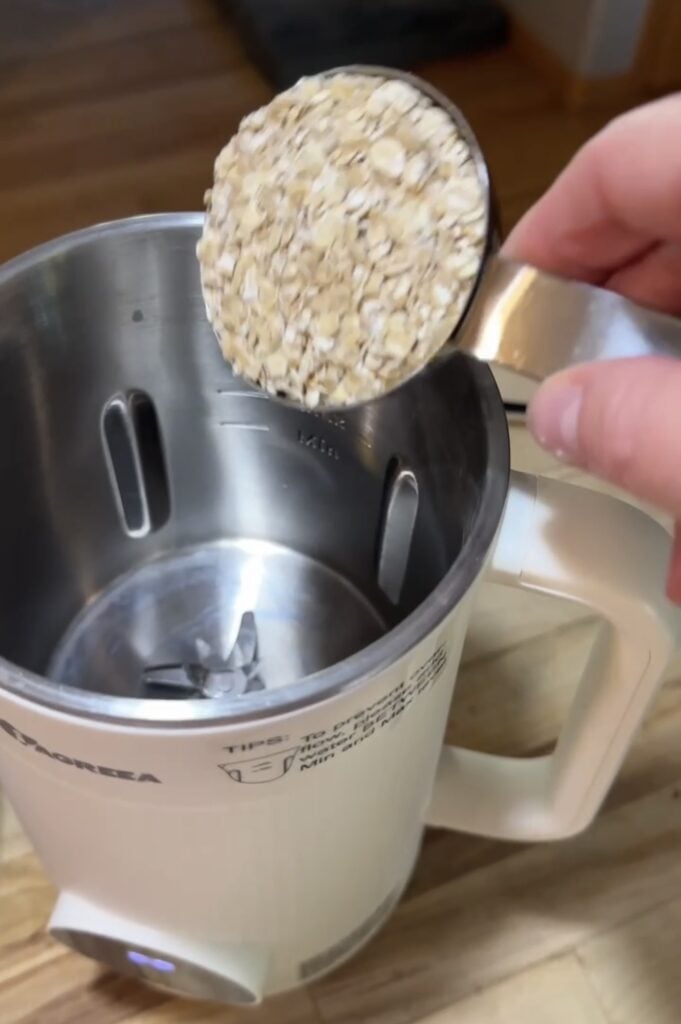 Rolled oats being added in a metal measuring cup to the stainless steel interior of the Iagreea nut milk maker machine, featured on slowestuff.com.