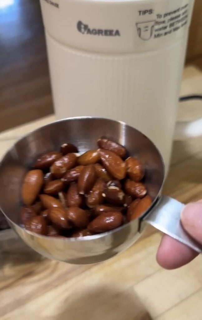 Close-up of soaked almonds measured in a metal scoop in front of the white Iagreea nut milk maker machine, featured on slowestuff.com.