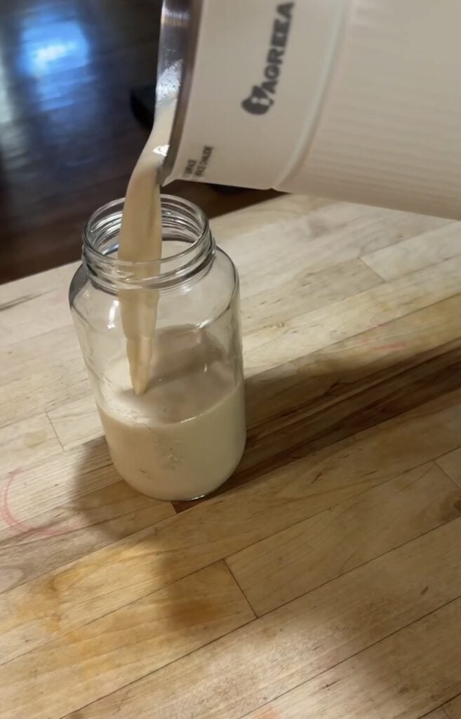Fresh homemade oat milk pouring directly from the white Iagreea nut milk maker machine into a clear glass jar, featured on slowestuff.com.