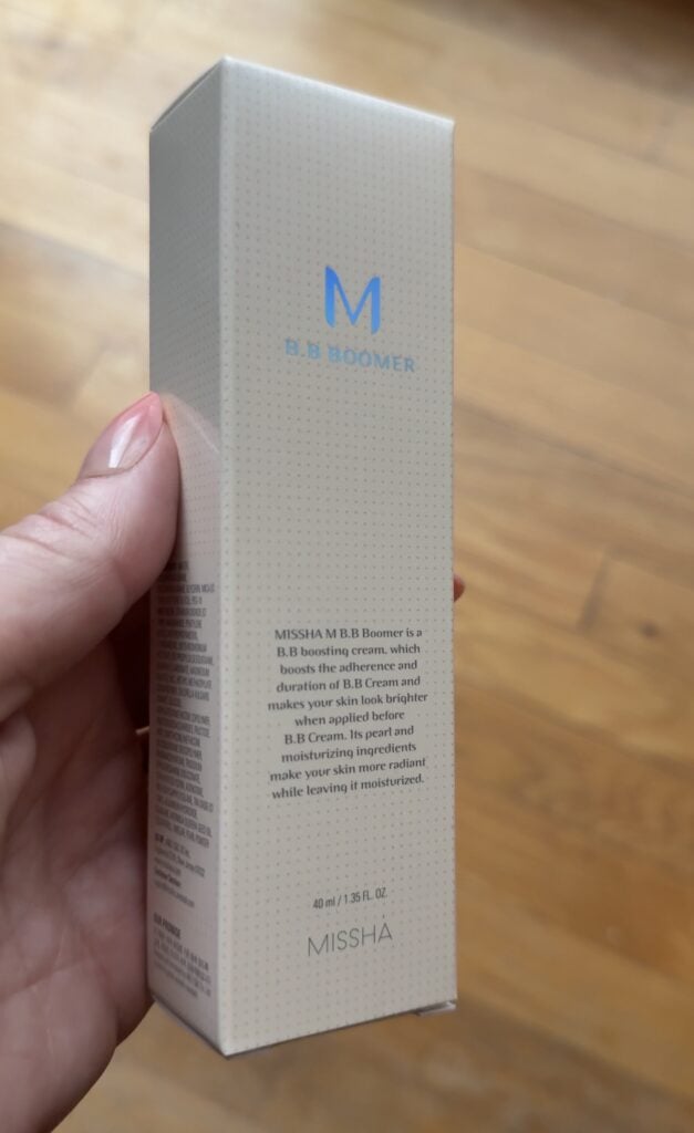 Hand holding the Missha M BB Boomer primer box on a wood floor background.