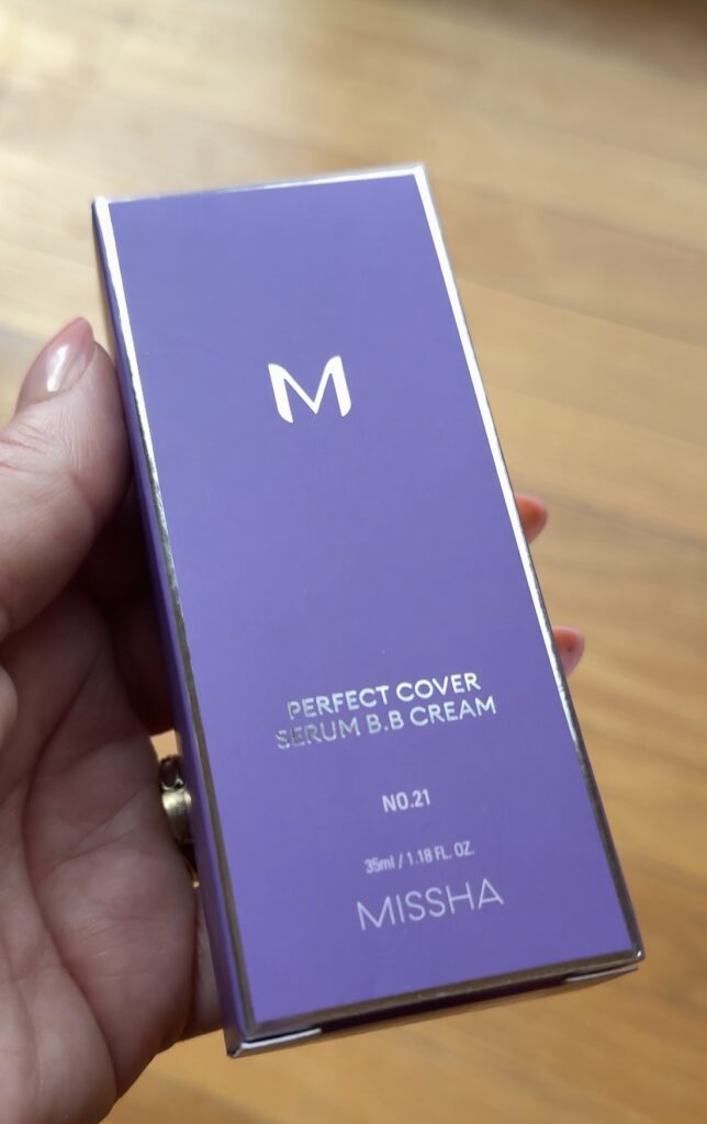 Hand holding the purple Missha Perfect Cover Serum BB Cream box in shade No. 21 on a wood floor background.