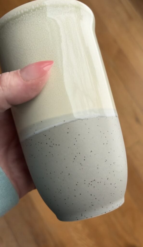 Close-up of a Nova Ceramics travel mug in a soft cream and speckled gray design, held in hand to show the smooth ceramic finish and curved shape.