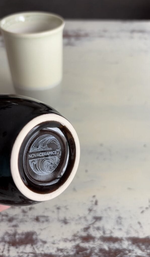 Bottom of a glossy dark Nova Ceramics travel mug showing the Nova Ceramics logo on the base, with the cream and gray mug blurred in the background.