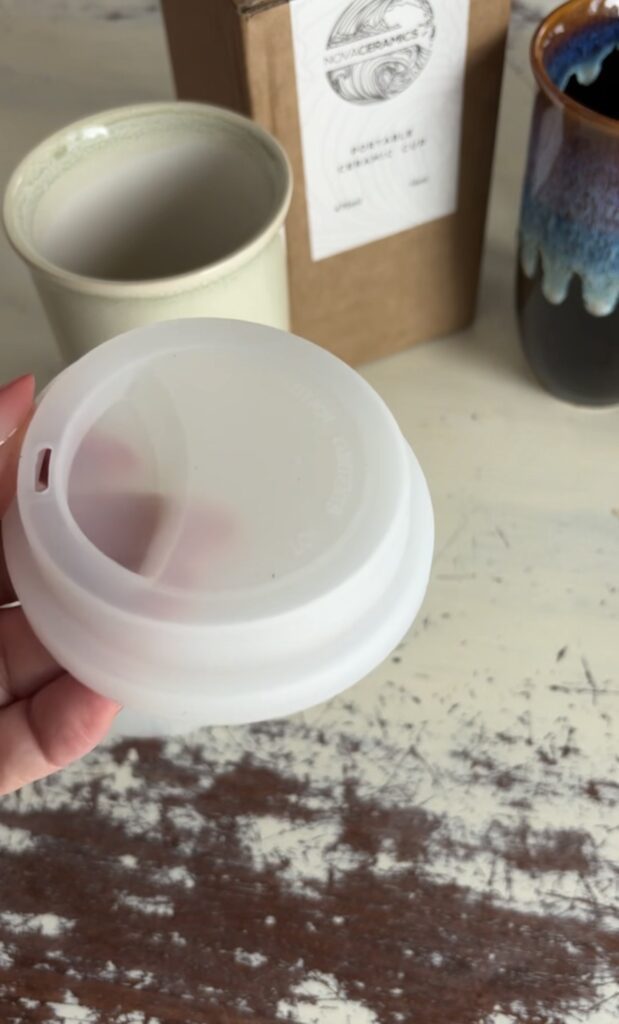 Frosted white silicone lid for a Nova Ceramics travel mug held in front of the ceramic mugs and branded packaging.