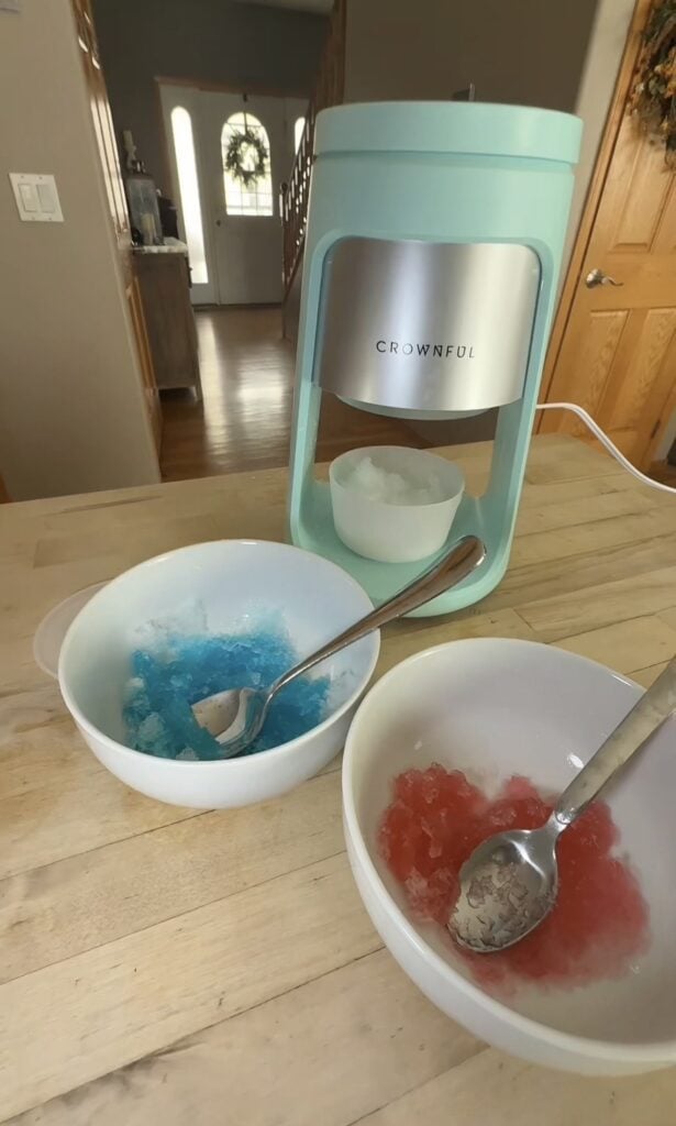 Two homemade snow cones with blue and red syrup in front of the Crownful shave ice machine.