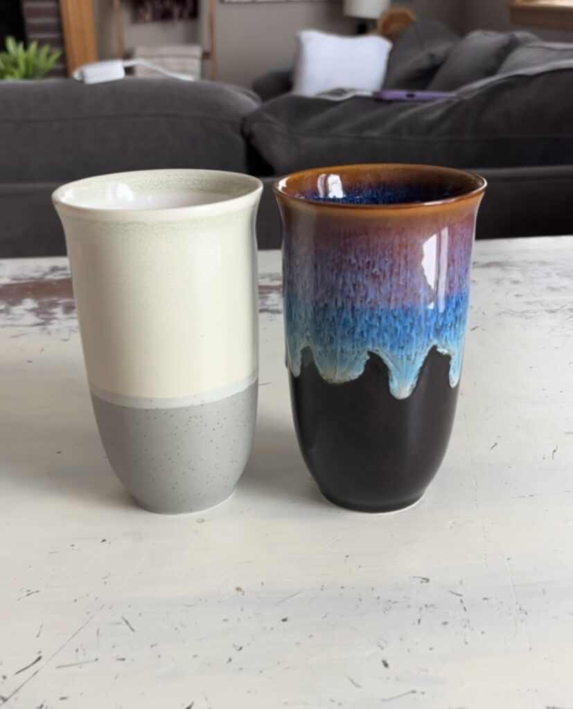 Two Nova Ceramics travel mugs standing side by side on a white distressed table, featuring one speckled cream and gray mug and one glossy black mug with blue and brown drip glaze.
