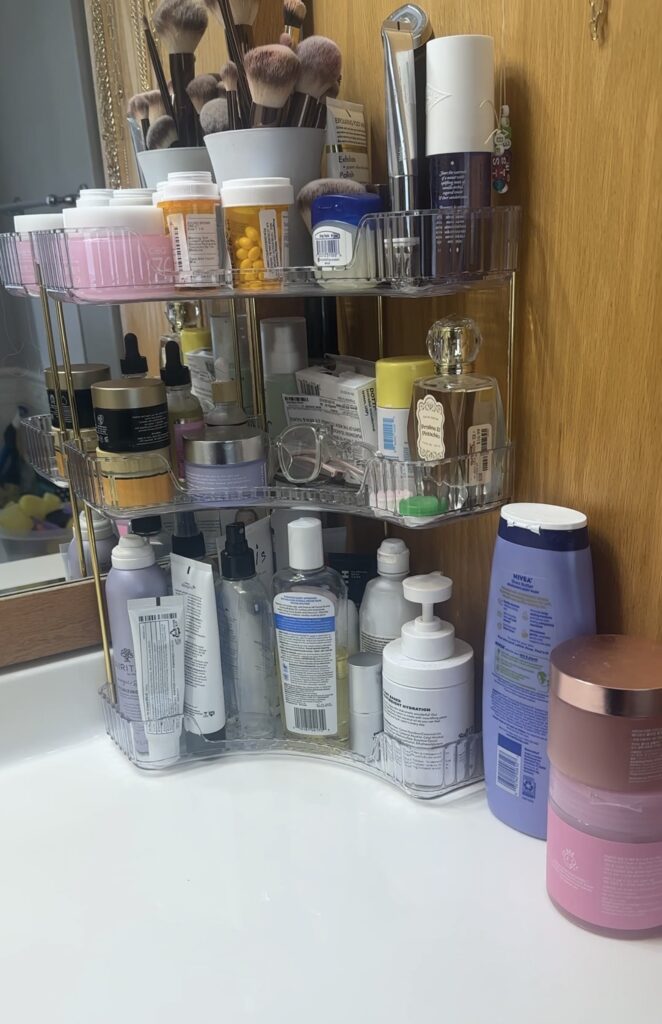 Bathroom countertop organizer shelf in clear acrylic and gold metal, used to store skincare, makeup, vitamins, perfume, and beauty products on a vanity.