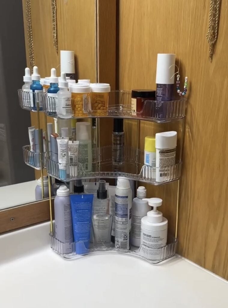 Corner bathroom countertop storage shelf with clear tiers and gold frame, organizing skincare, makeup, toiletries, and daily beauty essentials.