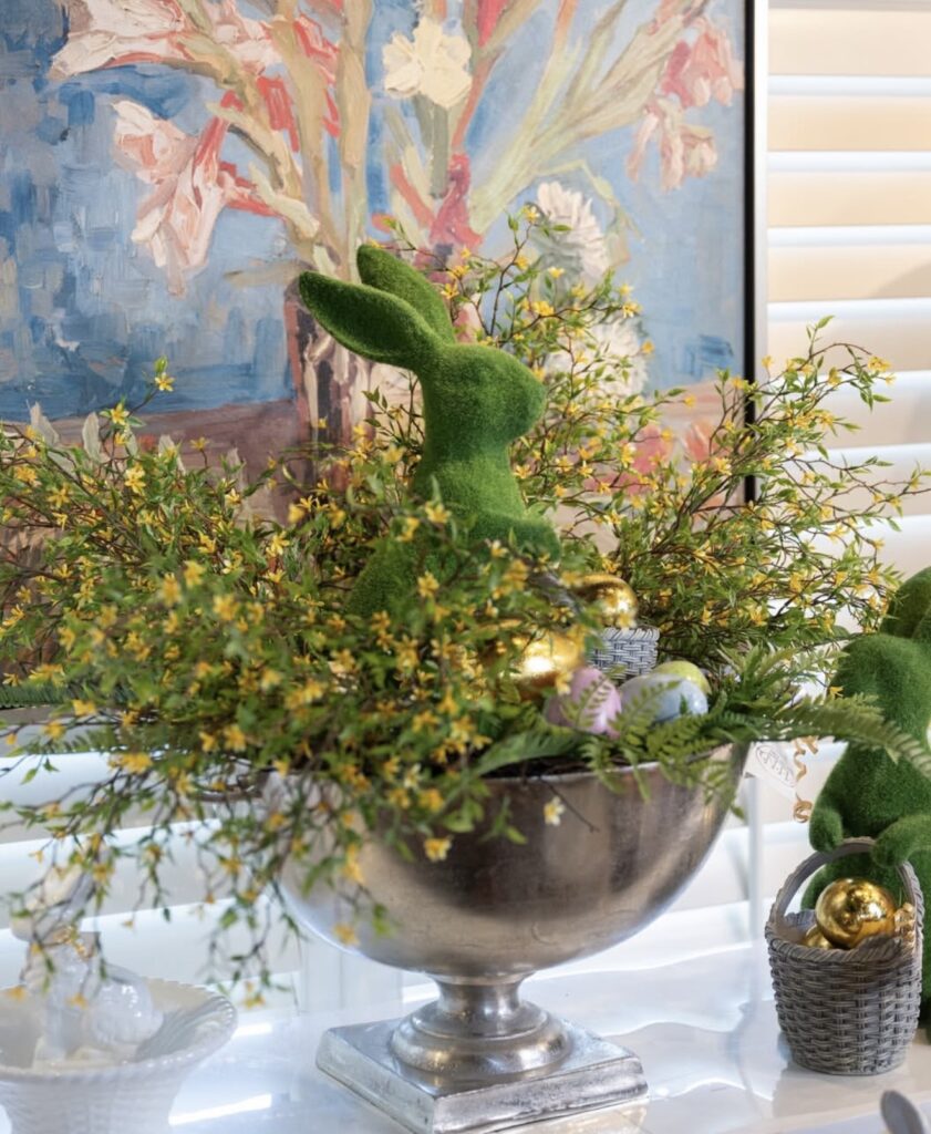 Easter centerpiece with a moss bunny, yellow flowering branches, pastel eggs, and gold eggs arranged in a silver urn in front of floral artwork.