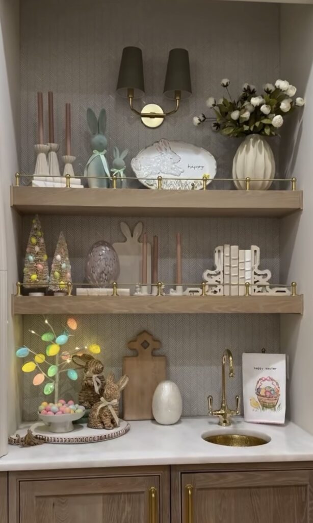 Elegant Easter kitchen shelf decor with pastel eggs, woven bunny figures, blush candles, spring florals, and a small gold faucet in a styled niche.