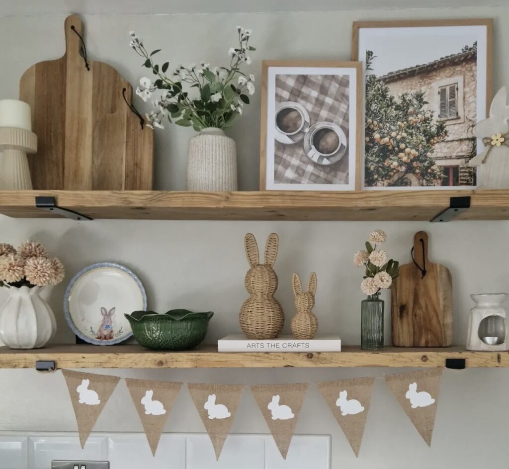Neutral Easter open shelf decor with wicker bunny figures, framed art, flowers, cutting boards, and bunny bunting in a soft farmhouse kitchen.