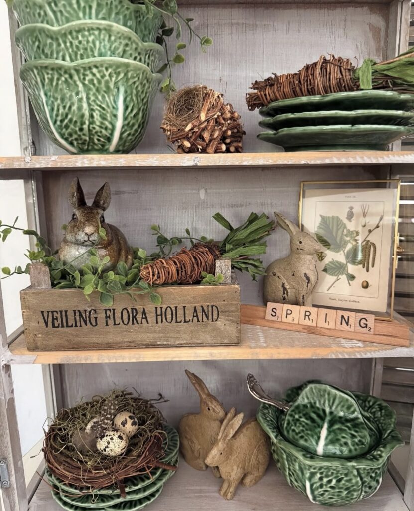 Earthy Easter kitchen shelf decor with cabbage plates, rustic bunny figures, woven carrots, bird nests, greenery, and warm vintage-inspired spring styling.