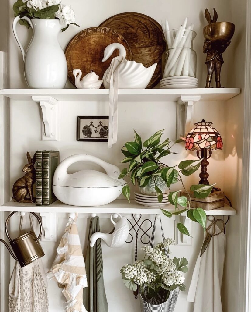Elegant Easter kitchen shelf decor with white swan dishes, stacked wooden cutting boards, a white pitcher of flowers, brass rabbit accents, a stained glass lamp, and hanging spring linens.