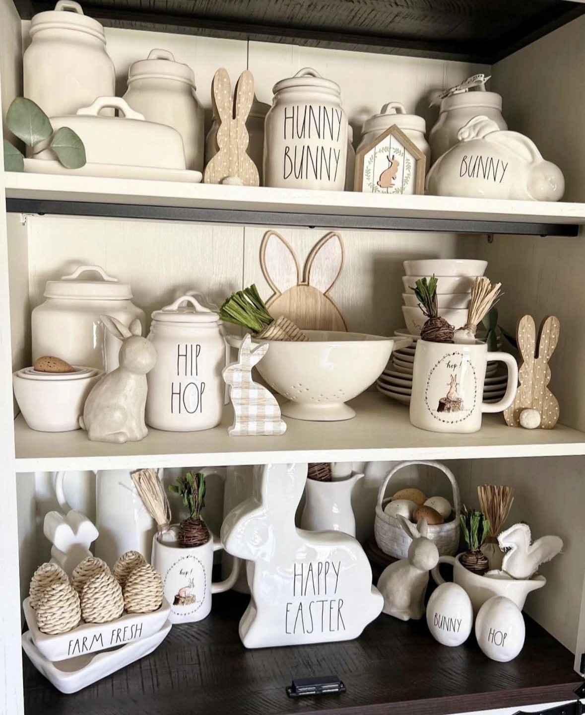 Rae Dunn Easter kitchen hutch decor with bunny canisters, ceramic rabbits, egg accents, mugs, and white spring kitchenware styled on dark shelves.