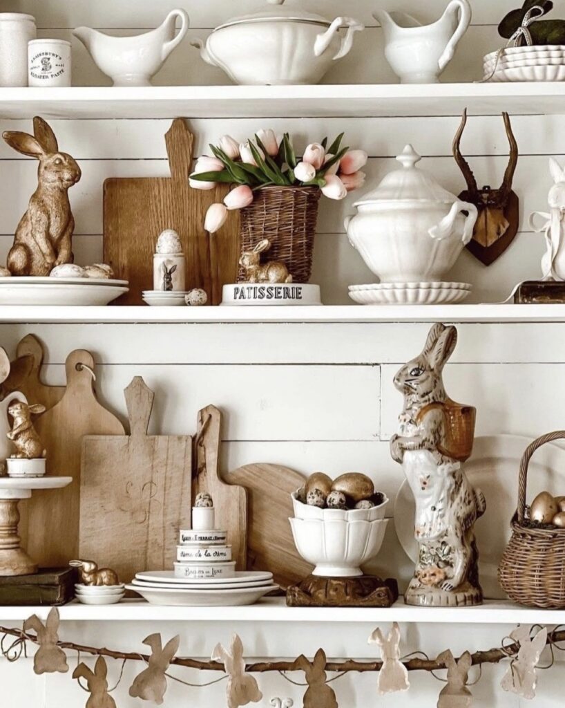 Vintage Easter kitchen shelf decor with brass rabbit figurines, pink tulips, white tureens, wood cutting boards, egg accents, and a bunny garland across open shelves.