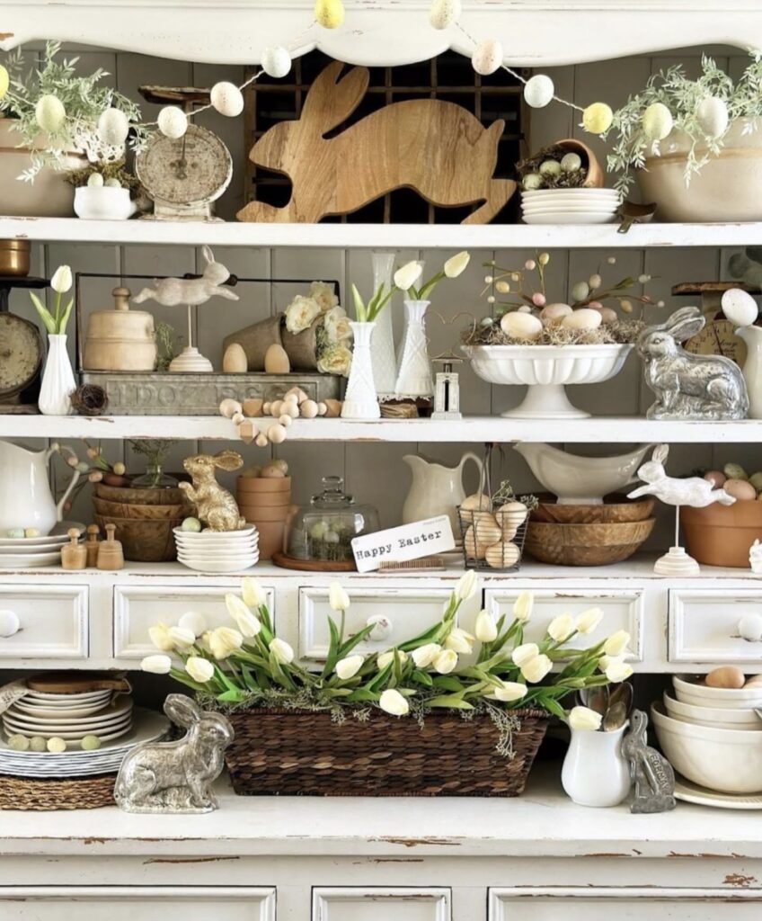 Rustic Easter hutch decor with white tulips, wooden bunny cutouts, silver rabbit figurines, pastel egg garland, white ironstone, and layered vintage spring styling.