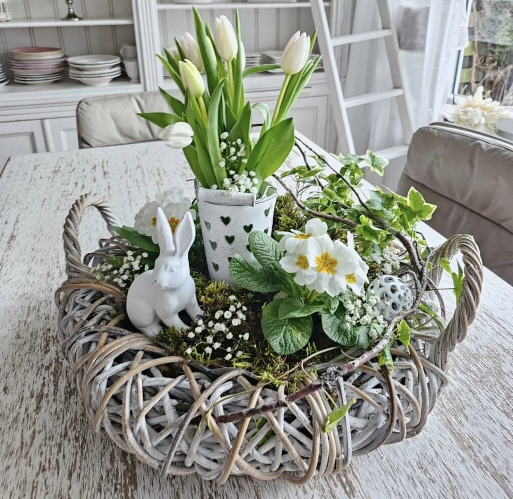 Fresh Easter table centerpiece with white tulips, primroses, moss, ivy, and a white ceramic bunny arranged in a woven basket tray.