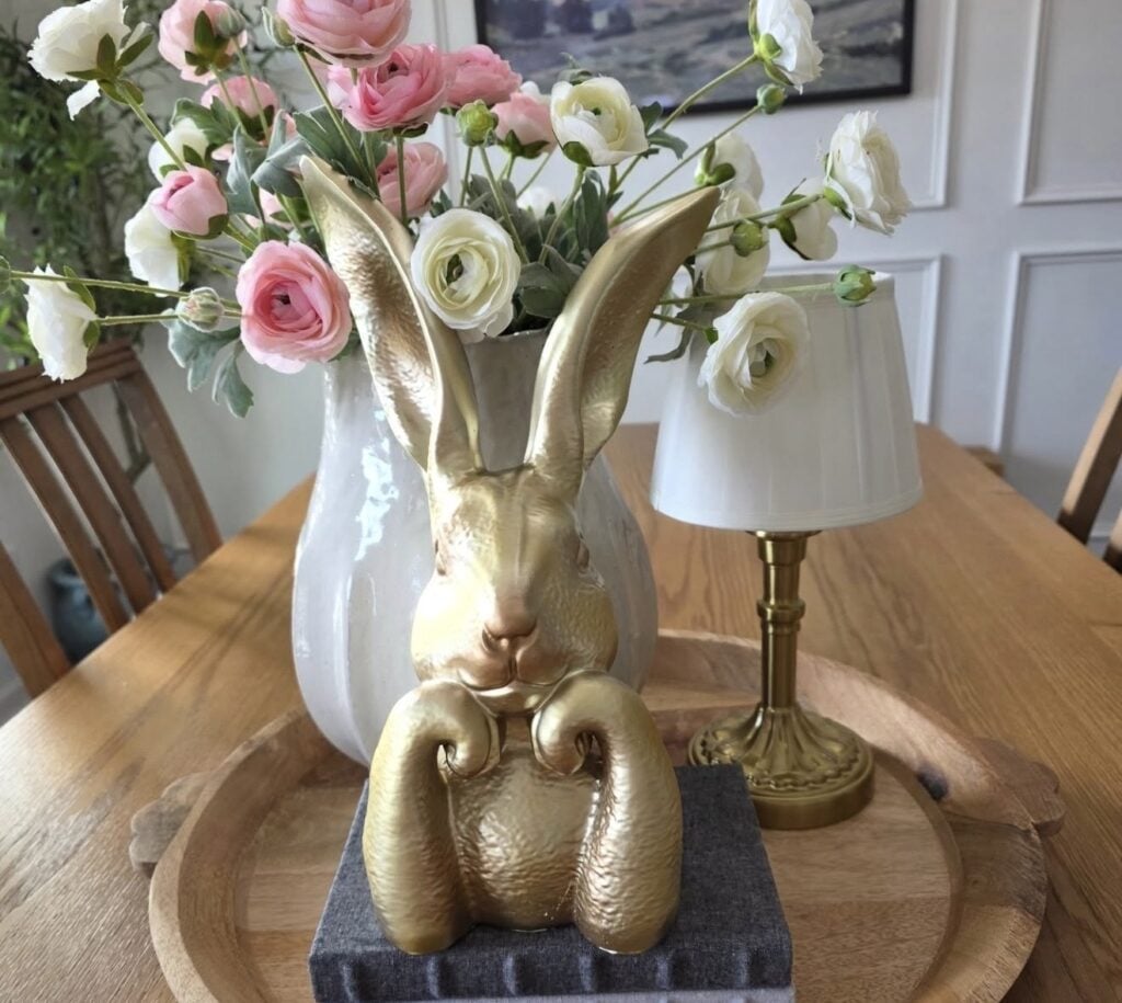 Elegant Easter table decor with a gold bunny statue, pink and white ranunculus in a white vase, and a small gold lamp on a wooden tray.