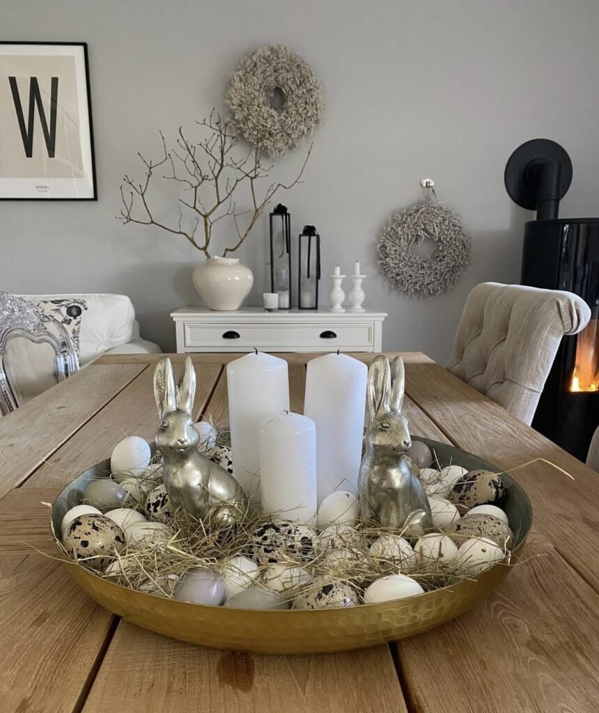 Neutral Easter dining table centerpiece with gold bunny figurines, white pillar candles, straw nests, and speckled eggs in a hammered gold bowl.