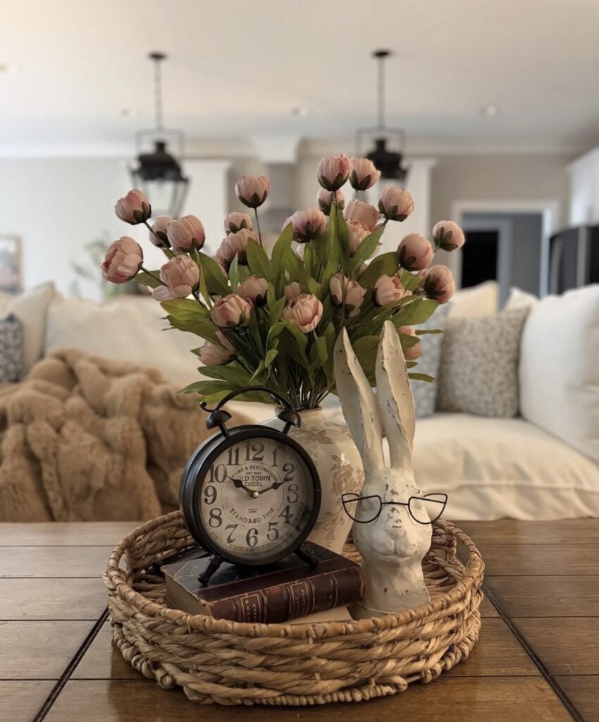 Cozy Easter coffee table decor with pink tulips, a rabbit bust wearing glasses, a vintage clock, and stacked books in a woven tray.