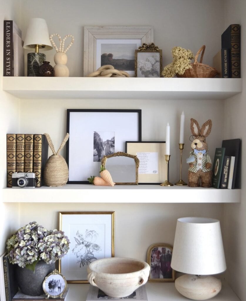 Neutral Easter shelf decor with framed art, vintage books, a wicker basket, bunny ear accents, candlesticks, a dressed rabbit figure, and soft cottage-style spring styling.