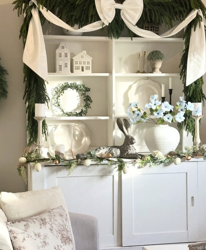 Elegant Easter shelf decor with greenery garland, a large white bow, ceramic houses, white platters, candlesticks, blue flowers, and a silver bunny centerpiece.