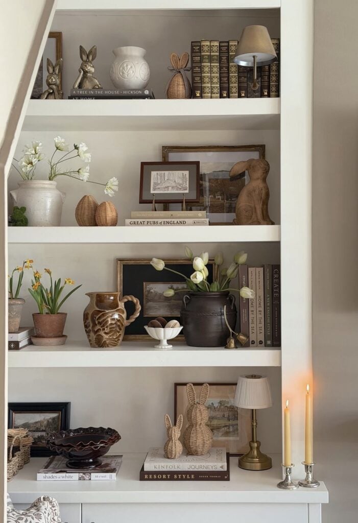 Neutral built-in Easter shelf decor with wicker bunny figurines, tulips, pottery, framed landscape art, vintage books, and brass candlesticks in a cozy living room.