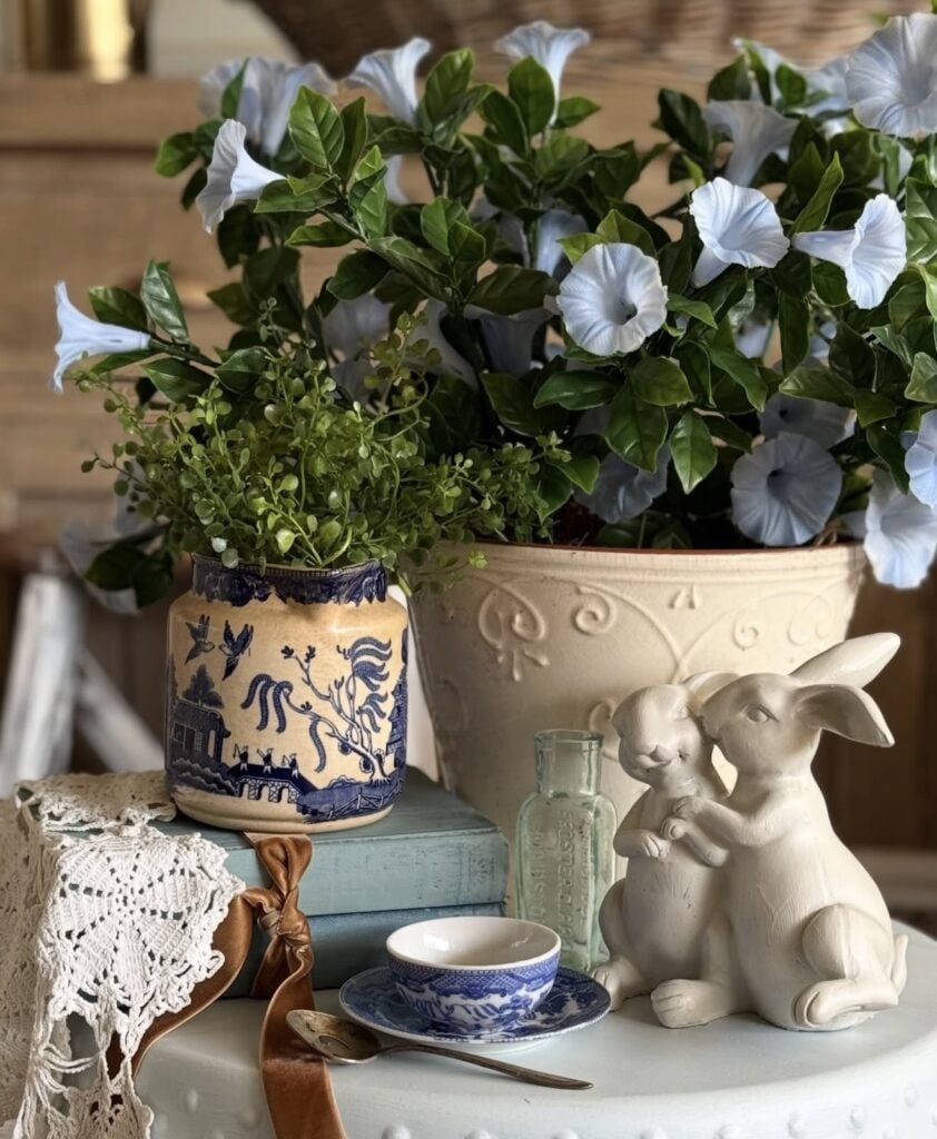 Blue and white Easter side table decor with a ceramic bunny couple, chinoiserie vase, lace details, blue teacup, and white flowering plant in a stone planter.