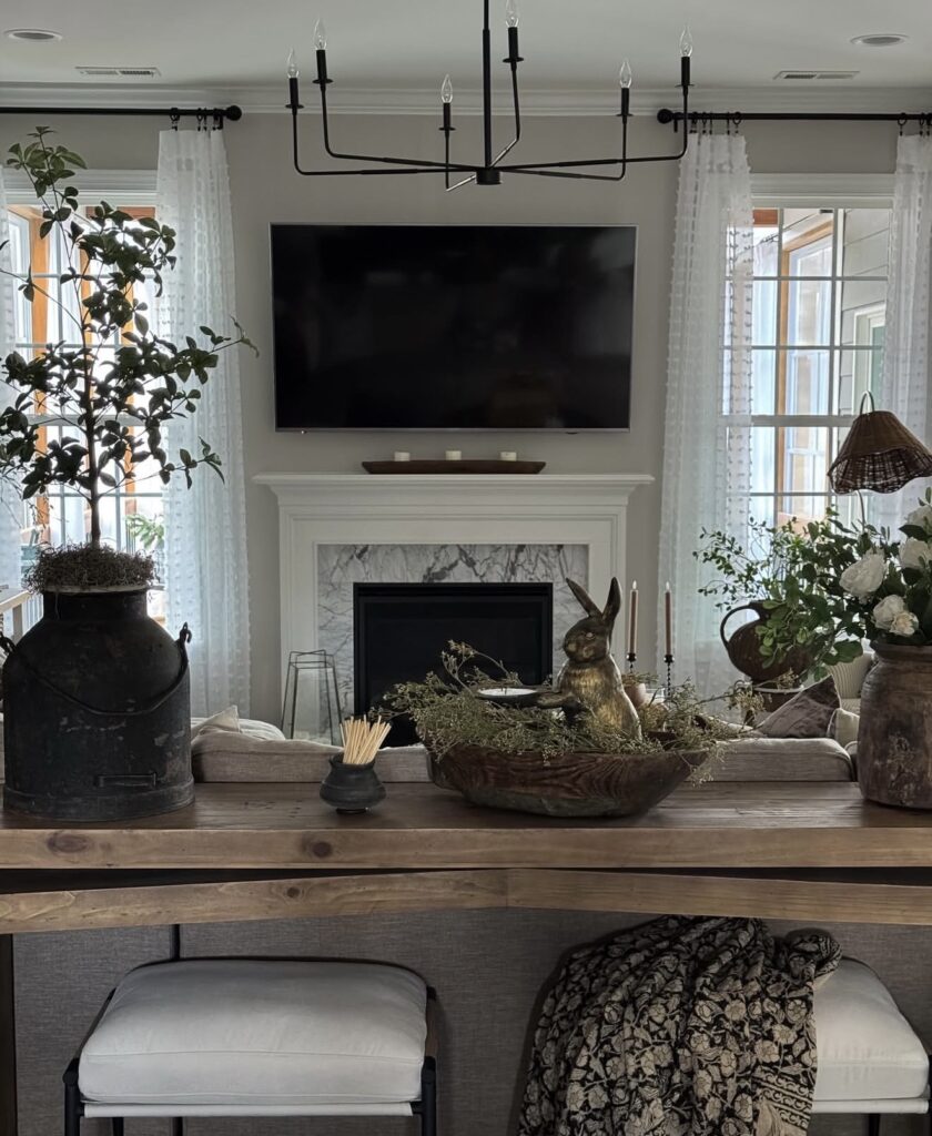 Neutral Easter console table decor with a black milk can, rustic wooden bowl, brass bunny figure, candles, and soft greenery in a bright living room.