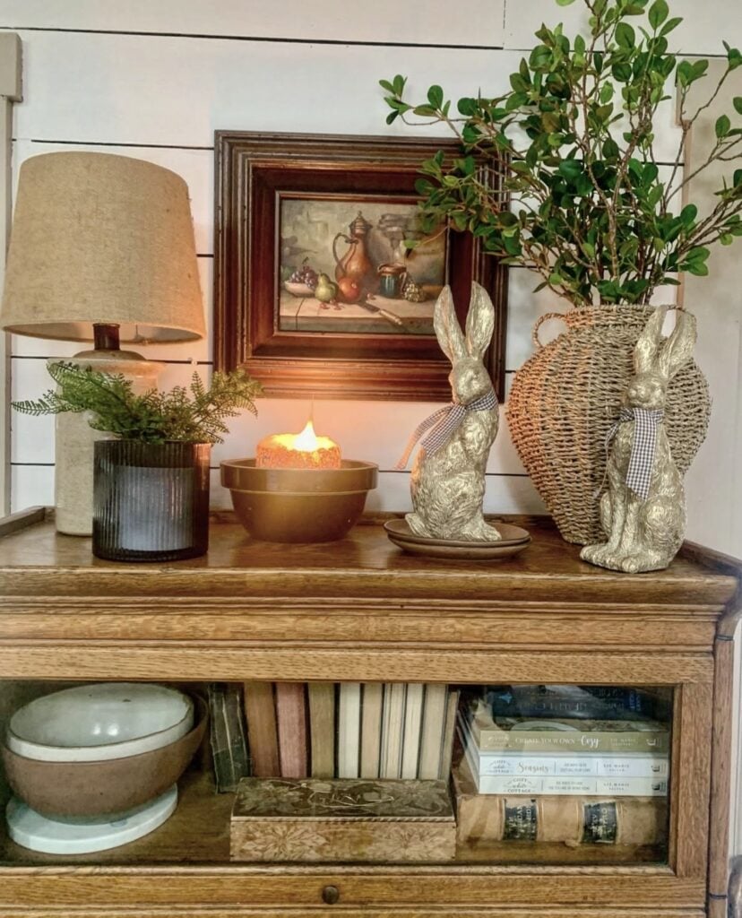 Cozy Easter table vignette with gold rabbit figurines, a woven vase, lamp, candle, greenery, and vintage art on a warm wood cabinet.