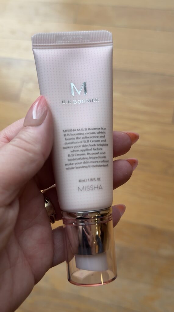 Hand holding the Missha M BB Boomer primer tube on a wood floor background.