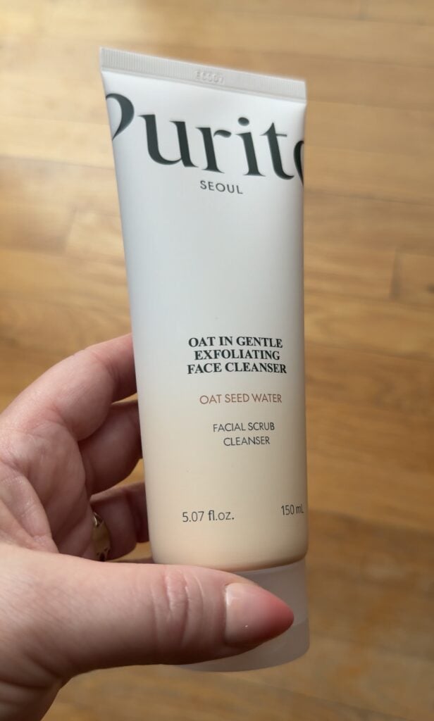Hand holding Purito Seoul Oat In Gentle Exfoliating Face Cleanser with oat seed water on a wood floor background.