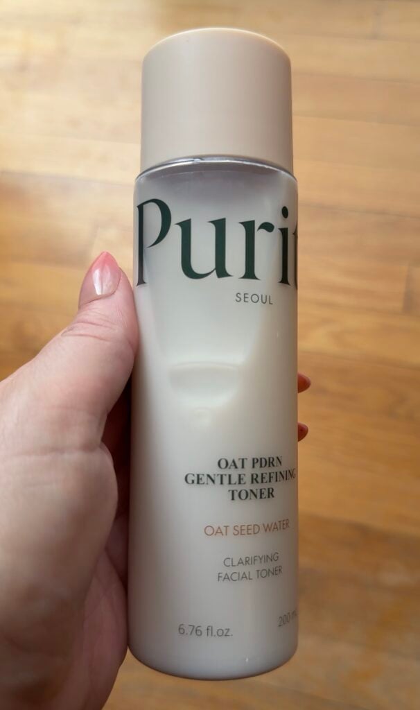 Hand holding Purito Seoul Oat PDRN Gentle Refining Toner with oat seed water on a wood floor background.