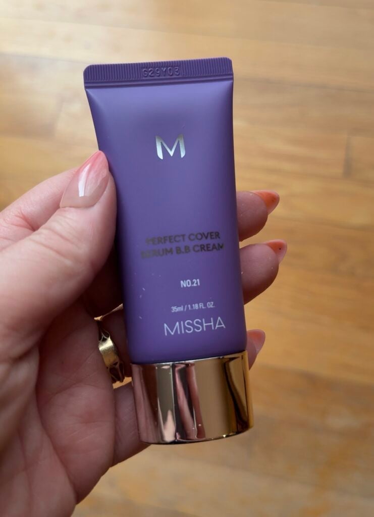 Hand holding the purple Missha Perfect Cover Serum BB Cream tube in shade No. 21 on a wood floor background.