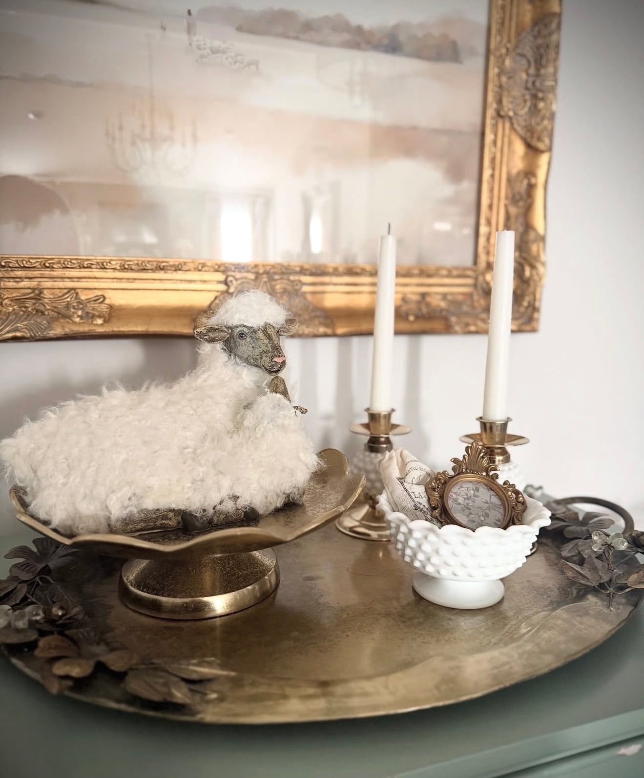 Elegant Easter tray decor with a fluffy lamb figure, brass candlesticks, a white milk glass bowl, and a gold framed mirror in soft neutral tones.