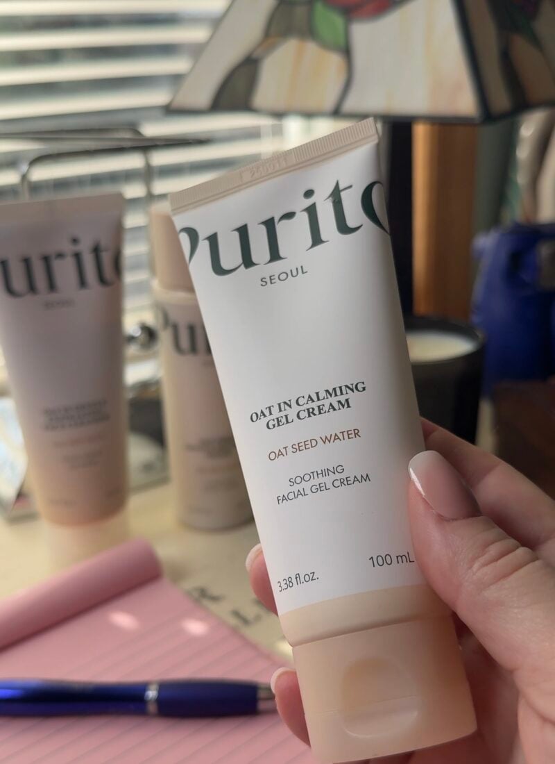 Hand holding Purito Seoul Oat In Calming Gel Cream with oat seed water in front of the full Purito skincare trio on a desk by a window.