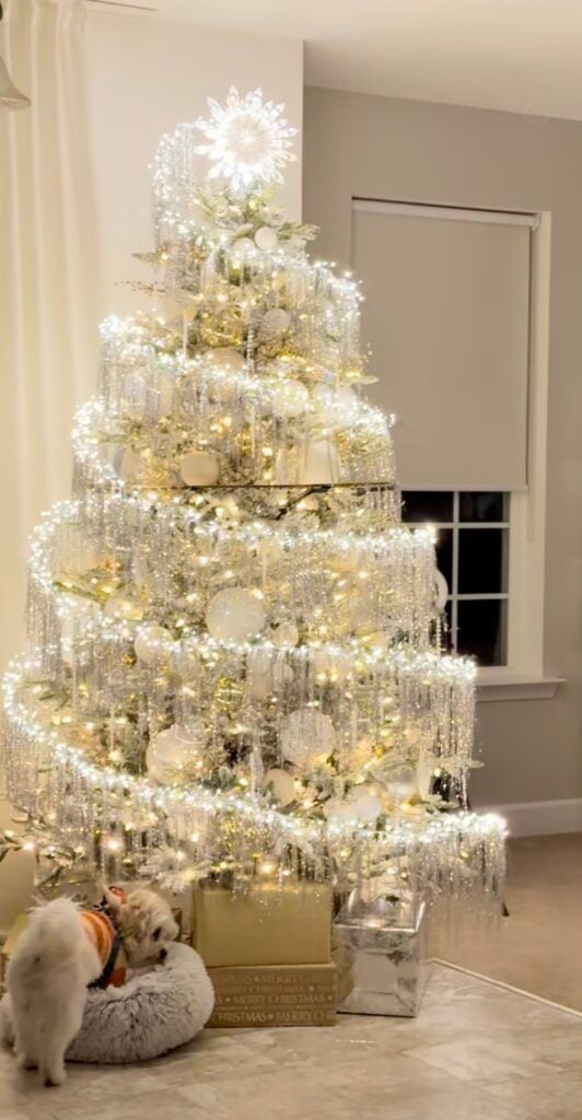 White Winter Solstice tree decorated with shimmering silver garland, warm white lights, oversized white ornaments, and a glowing snowflake tree topper.