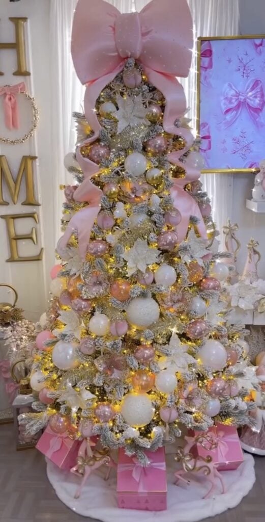 Flocked Christmas tree decorated with pink, white, and gold ornaments, white poinsettias, warm lights, and a giant pink bow topper.