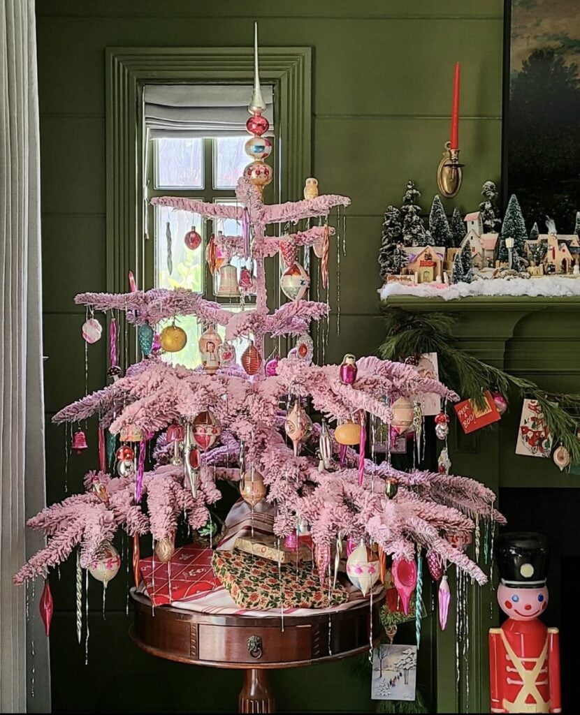 Sparse pink Christmas tree decorated with colorful vintage glass ornaments and icicles against a dark green wall and vintage holiday display.