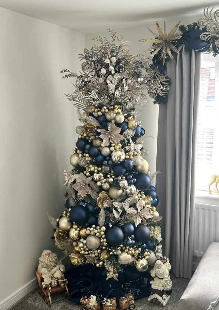 Full Christmas tree styled with blue ornaments, ribbon, decorative picks, and warm white lights for an elegant holiday look.