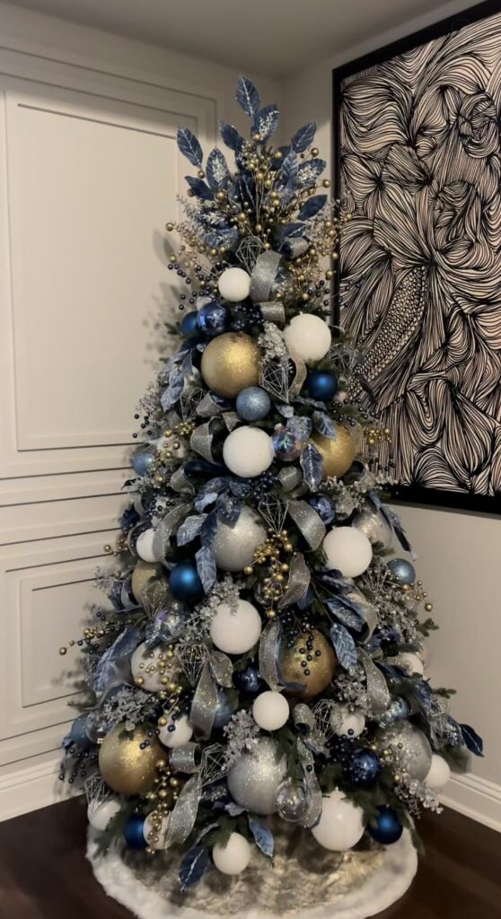 Blue Christmas tree decorated with gold and silver ornaments, metallic ribbon, reflective accents, and glowing white lights.