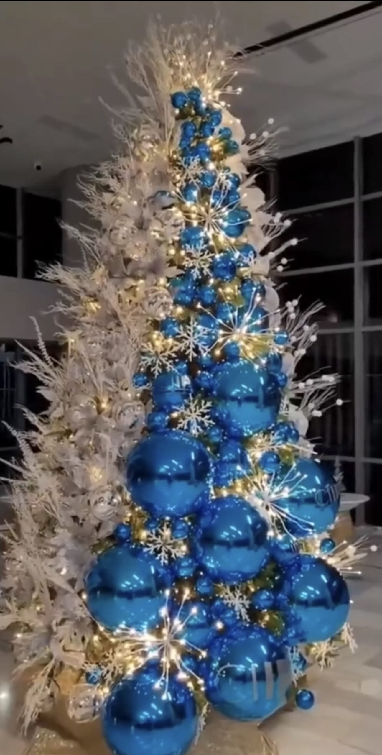 Christmas tree decorated with oversized blue ornaments, silver accents, large ribbon, and dramatic holiday decorations.