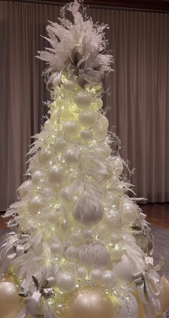 White Christmas tree decorated with snowy branches, white florals, reflective ornaments, delicate icicles, and warm glowing lights.