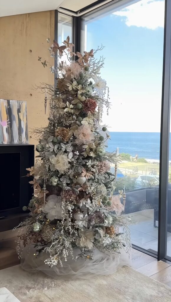 Christmas tree decorated with large florals, greenery, ornaments, ribbon, and botanical picks for a dramatic holiday display.