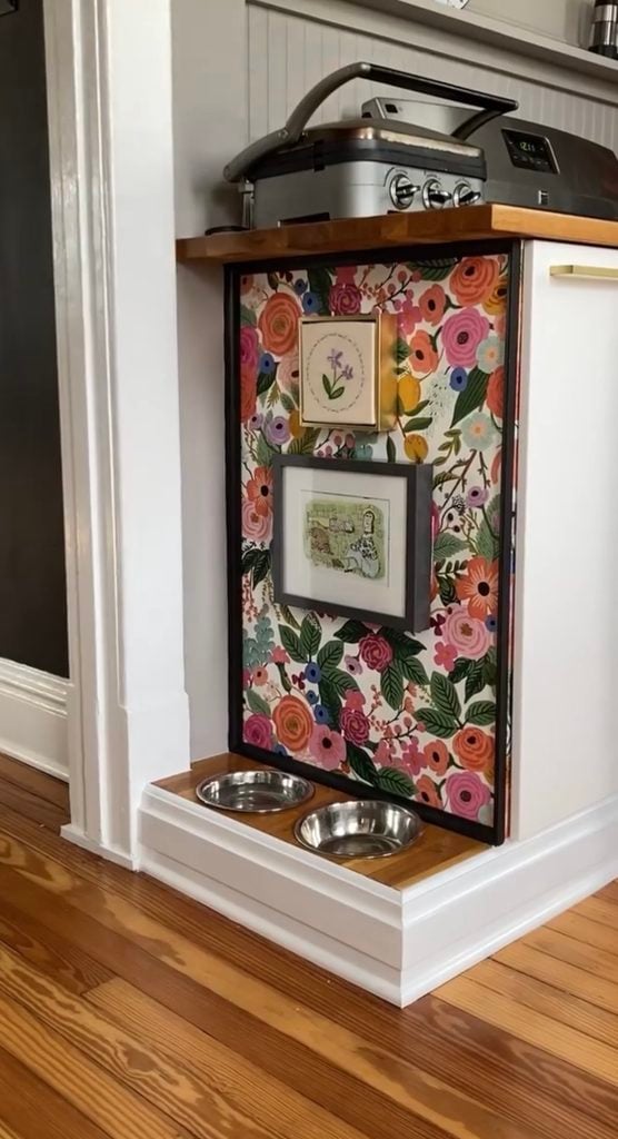Built-in dog feeding station in a kitchen with colorful floral wallpaper and two framed pieces of artwork above stainless steel bowls.