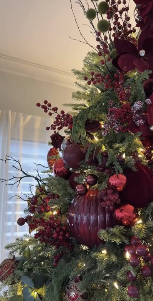Burgundy floral Christmas tree decorated with deep red flowers, berries, ornaments, greenery, and warm white lights.