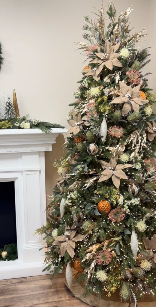 Christmas tree decorated with oversized fabric flowers, ornaments, greenery, and layered floral accents.