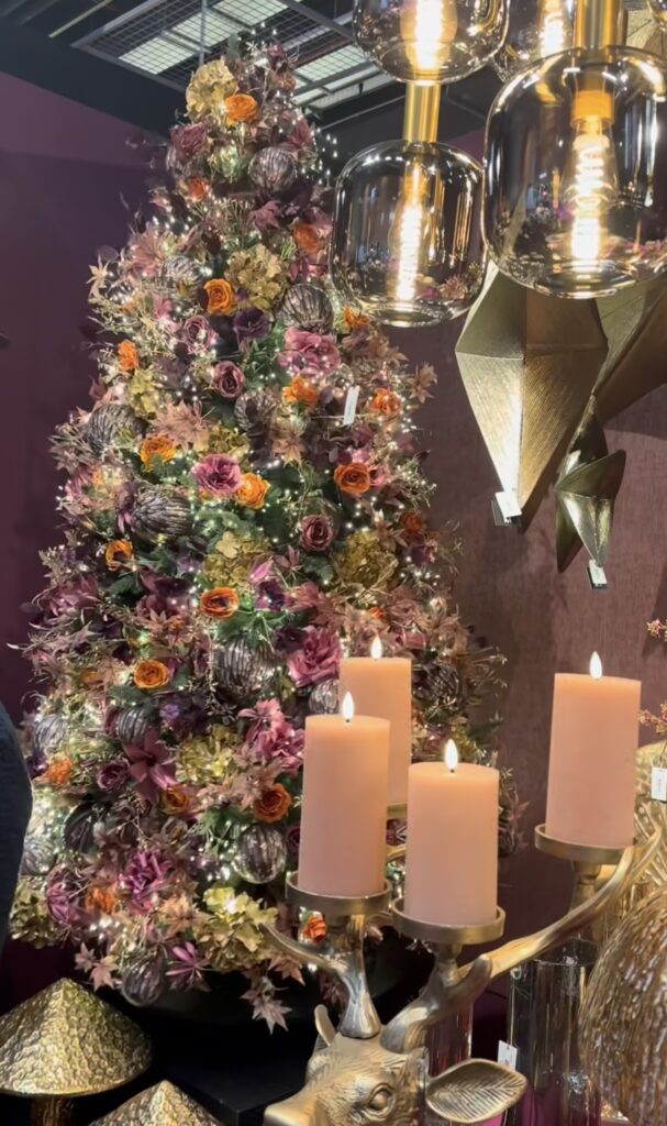 Lush floral Christmas tree covered with burgundy, mauve, orange, and blush roses, hydrangeas, oversized metallic ornaments, greenery, and warm white lights.