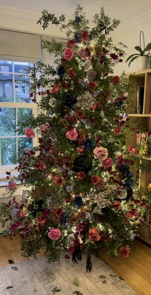 Christmas tree decorated with large flowers, layered greenery, ornaments, and botanical stems tucked throughout the branches.