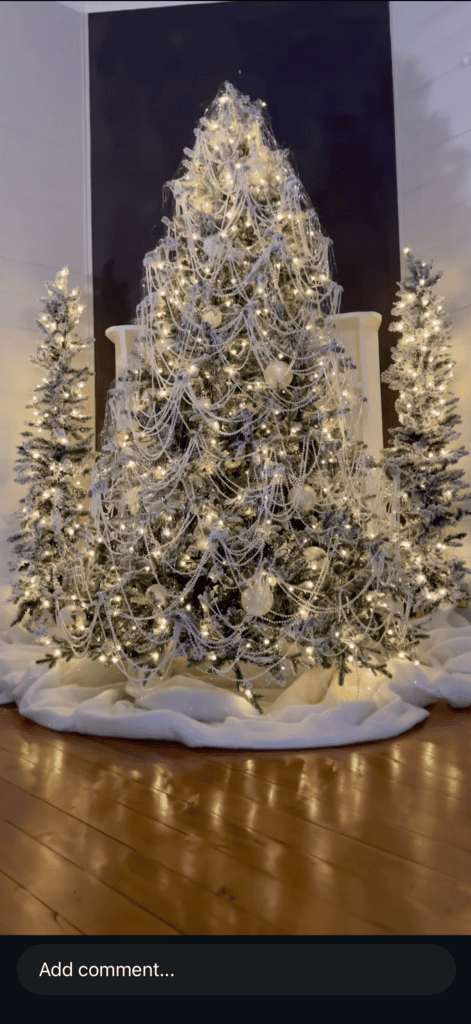 Green Christmas tree draped with layers of pearl garland, clear bead strands, white ornaments, and warm lights, flanked by two smaller lit trees.